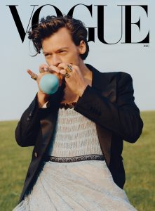 A portrait of Harry Styles blowing a balloon while wearing a black blazer and a grey dress. 