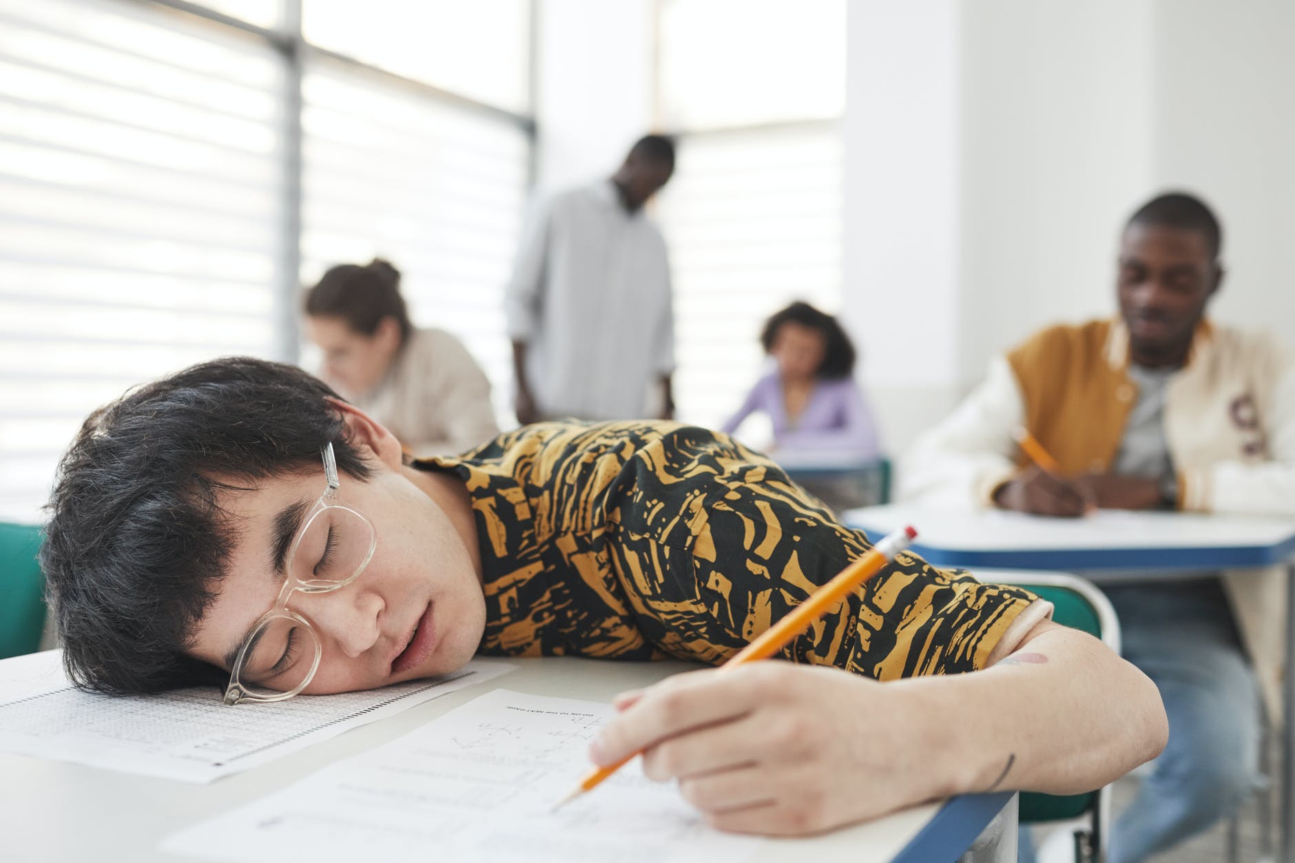 Photo of a person lying with their cheek and shoulder on the desk in front of them while they sit. Their eyes are closed and their hand is holding a pencil, poised to write, however they are asleep
