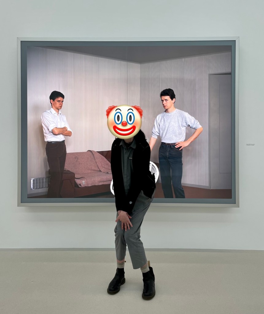 A person squatting in front of a large photograph. The photograph depicts to identical young men, standing around a chair and looking sternly at the camera. The person in front of the photograph is squatting as iff pretending to sit in the chair. They are wearing a gray uniform and a clown emoji obscures their face.