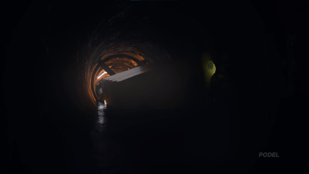 A screenshot from the YouTube video "SpongeBob Experiences Delta P and Dies Instantly". SpongeBob's upper body can be seen at the center of the frame, he's laying face down in a oil rigging pipe. The scene is dimly lit, light comes from 2 cracks in the pipe, one behind SpongeBob, faintly illuminating the surrounding orange rusty innards of the pipe, the oil, and the back of SpongeBob's head. The other source of light coming from the crack that SpongeBob's body occupies, where a small portion of is yellow body is visible.