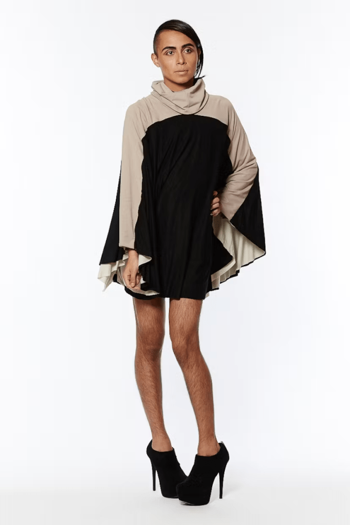 Person with Nude make up
unshaved legs 
wearing a black poncho with cowl turtle neck and arms are sewn in to make sleeves that are part of the poncho, the arms, shoulders and turtle neck are all a light beige making a single light beige stripe at the top of the garment
black boot heels 
white backdrop