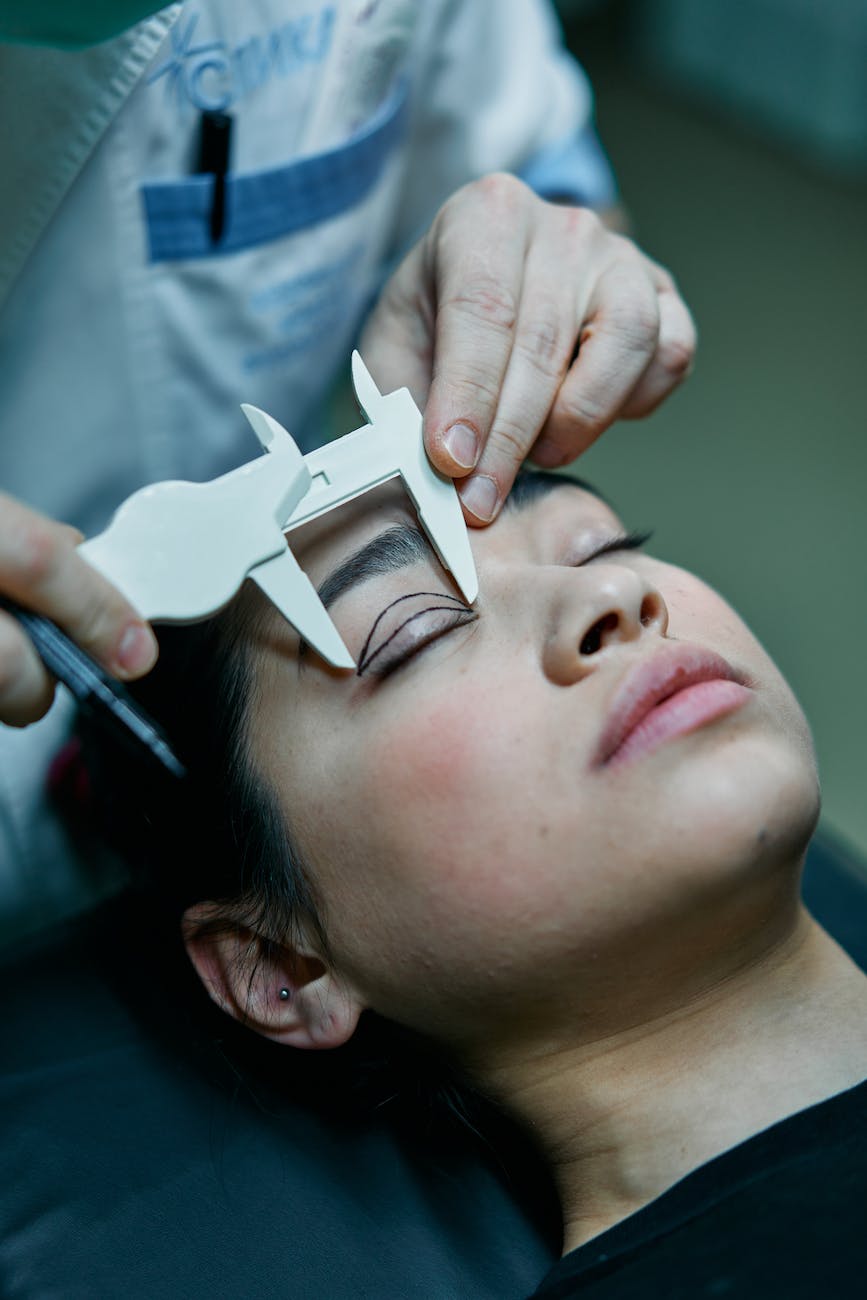 This image shows a woman laying with her eyes closed. There is a surgeon measuring and marking lines on her eyelid. She seems to be soon undergoing a plastic surgery procedure.