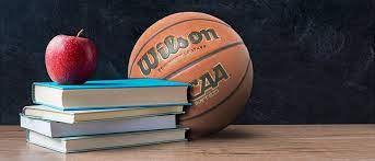 A stack of books with an apple on top and a basketball behind the stack. All three item are placed on a desk with a dark backround.