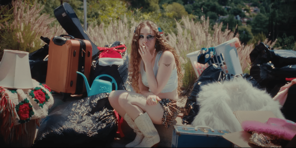 Chappell Roan: A woman with red curly hair with tear-streaked makeup, white tank top and cheetah print shorts, and white cowboy boots sitting on the curb smoking a cigarette. She is surrounded by disorganized piles of her belongings.