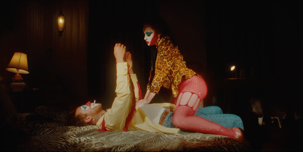 Chappell Roan and Clownie: A woman with red curly hair, clown makeup, cheetah print blazer and red fishnets straddling a man in clown makeup, yellow shirt, and blue jeans on a cheetah print bed. The room is dimly lit with a spotlight on Chappell Roan and Clownie.