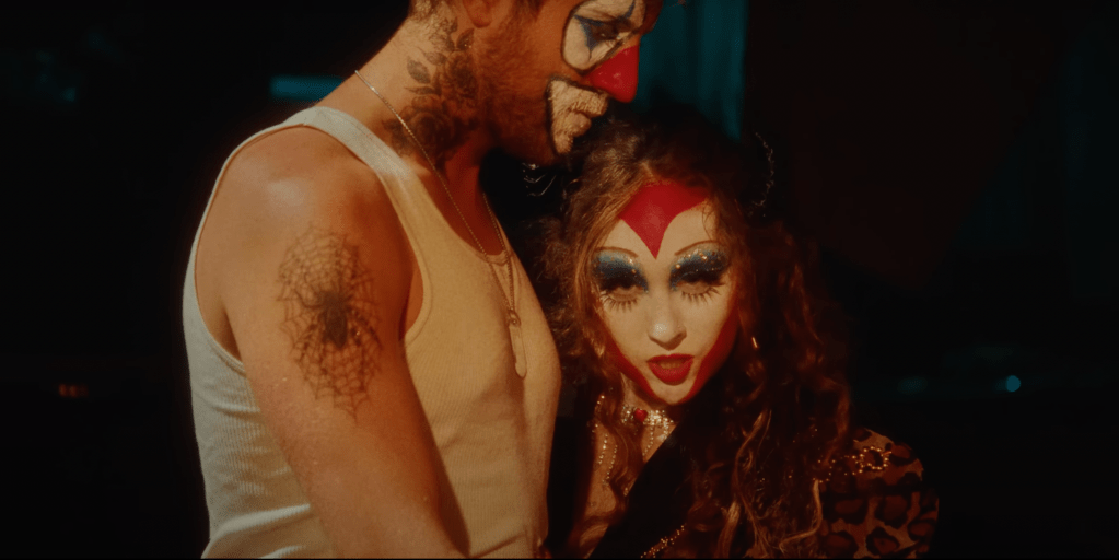 Chappell and Clownie: A woman with red curly hair, a cheetah print blazer and heart-shaped clown makeup is staring into the camera, while a man in clown makeup and a white undershirt is staring down blankly.