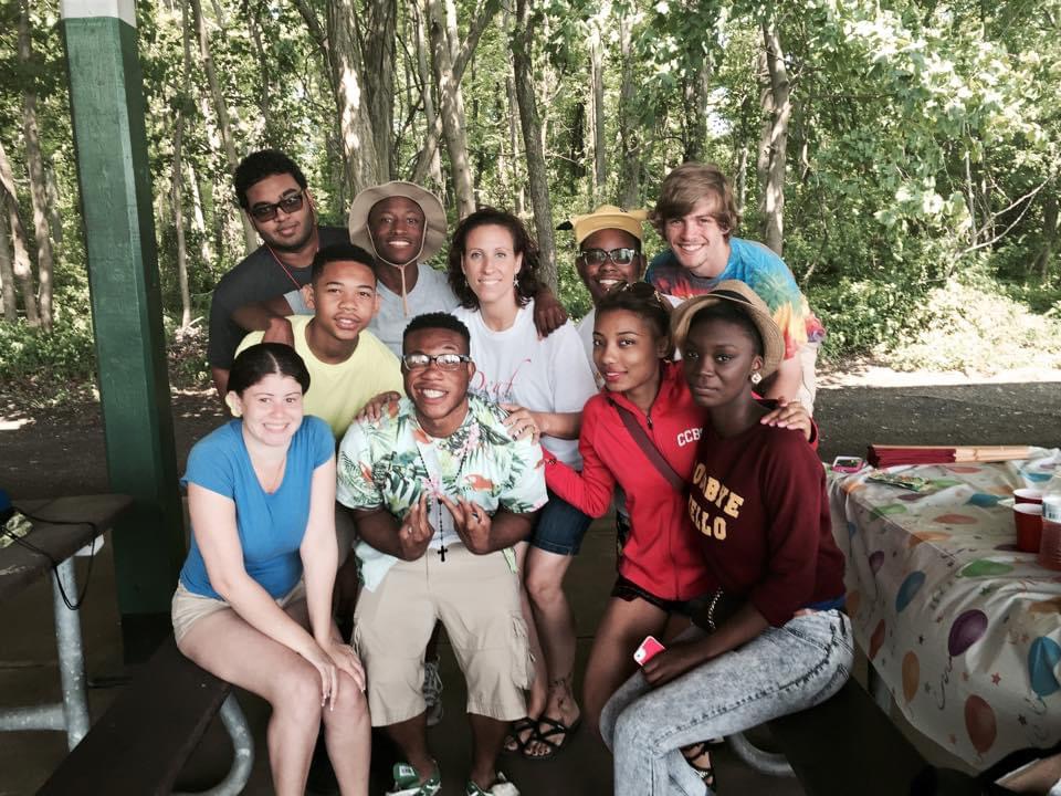 a group of students, some sitting and some standing, posing for a picture with their teacher under a pavilion on a bright and sunny day. 