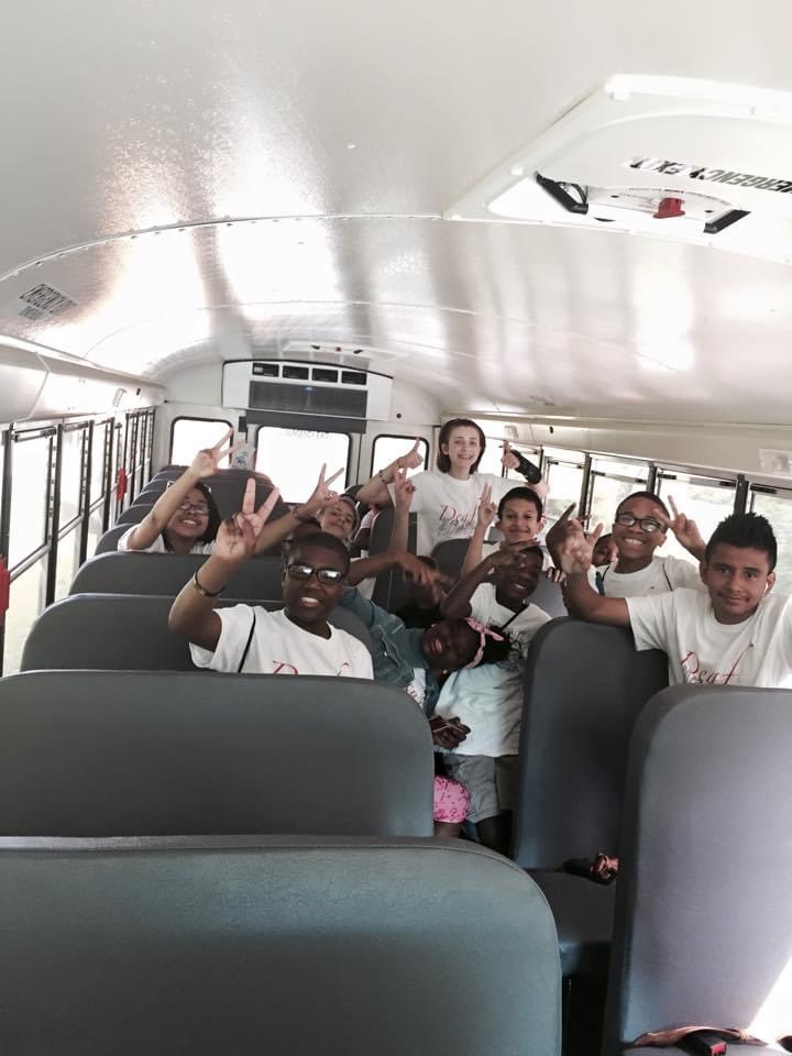 Group of smiling children on a school bus, wearing matching white t-shirts that say "Deaf Fest" in red text. All of the children are holding peace signs up with their hands while smiling at the camera. 