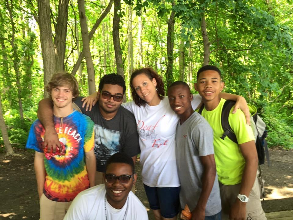 A group of students smiling for a picture with their teacher outdoors. The background is bright green with trees and greenery, with the sun peaking out of the trees. 
