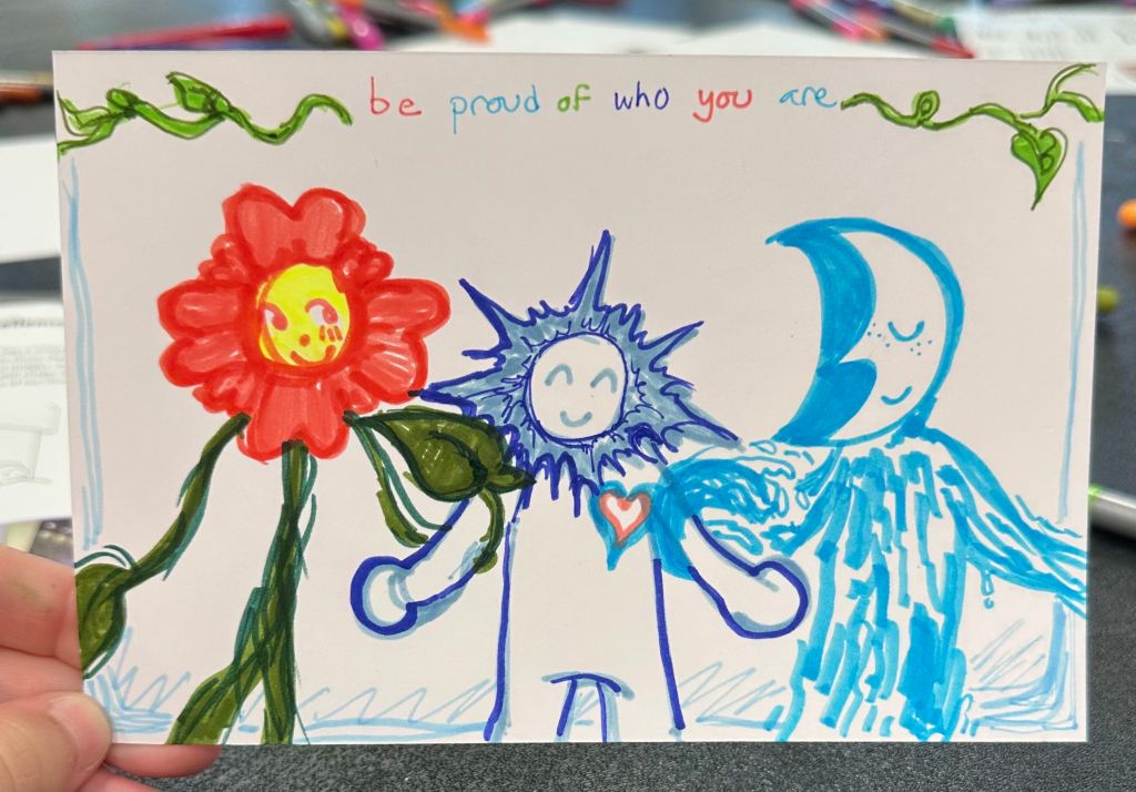 A white notecard with three figures. On the right stands a flower person with pink petals and their green vines are wrapped around the figure to the left of them, a simple figure with a star-like "mane" around their head. To the right of them is a figure with a crescent moon head and a body like water. The moon's arm is shaped like a wave and is intertwined with the middle figure. Above them are green vines and the text "be proud of who you are".